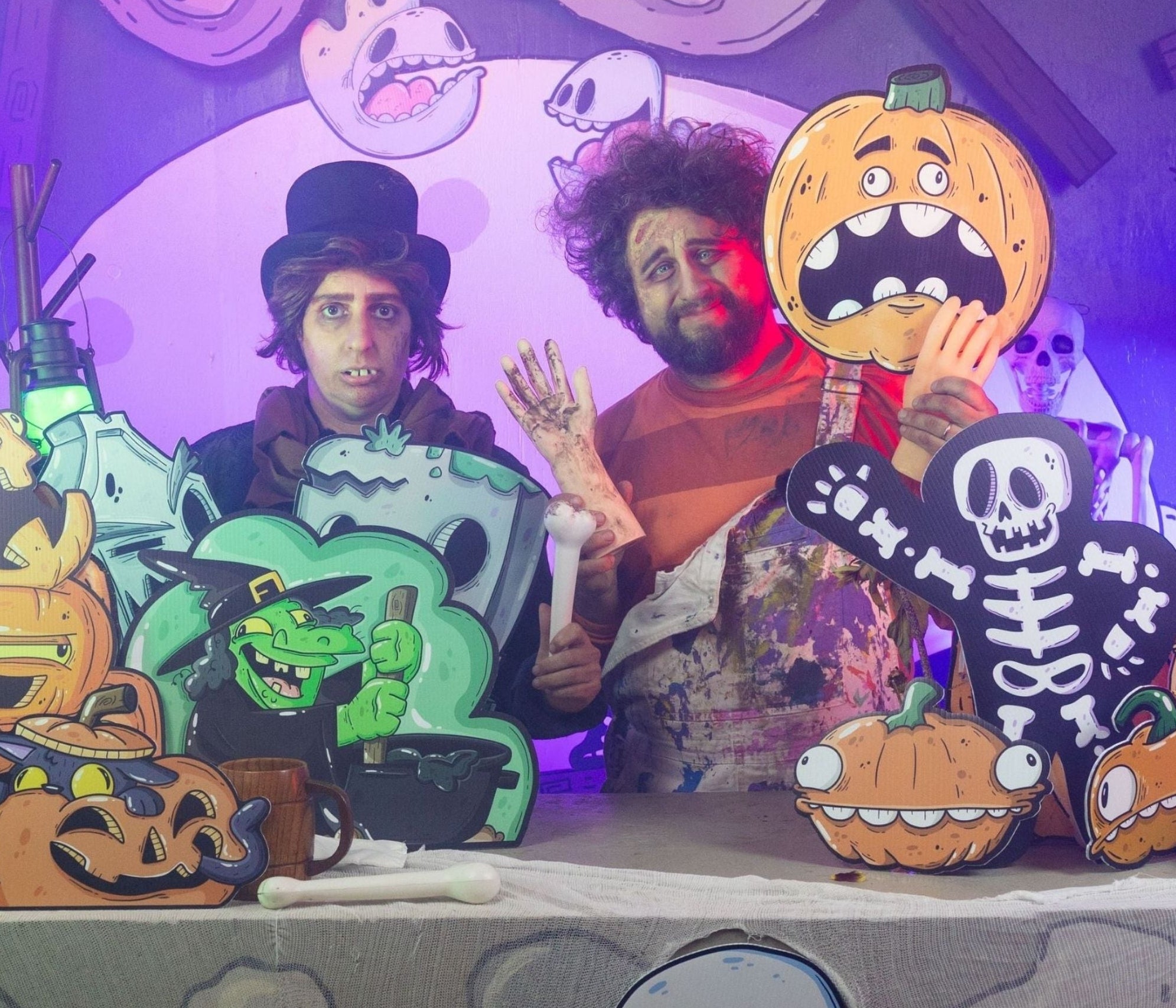 Two people dressed in halloween costumes standing behind a table with cartoon decorations including pumpkins, a witch, skeleton and ghosts.